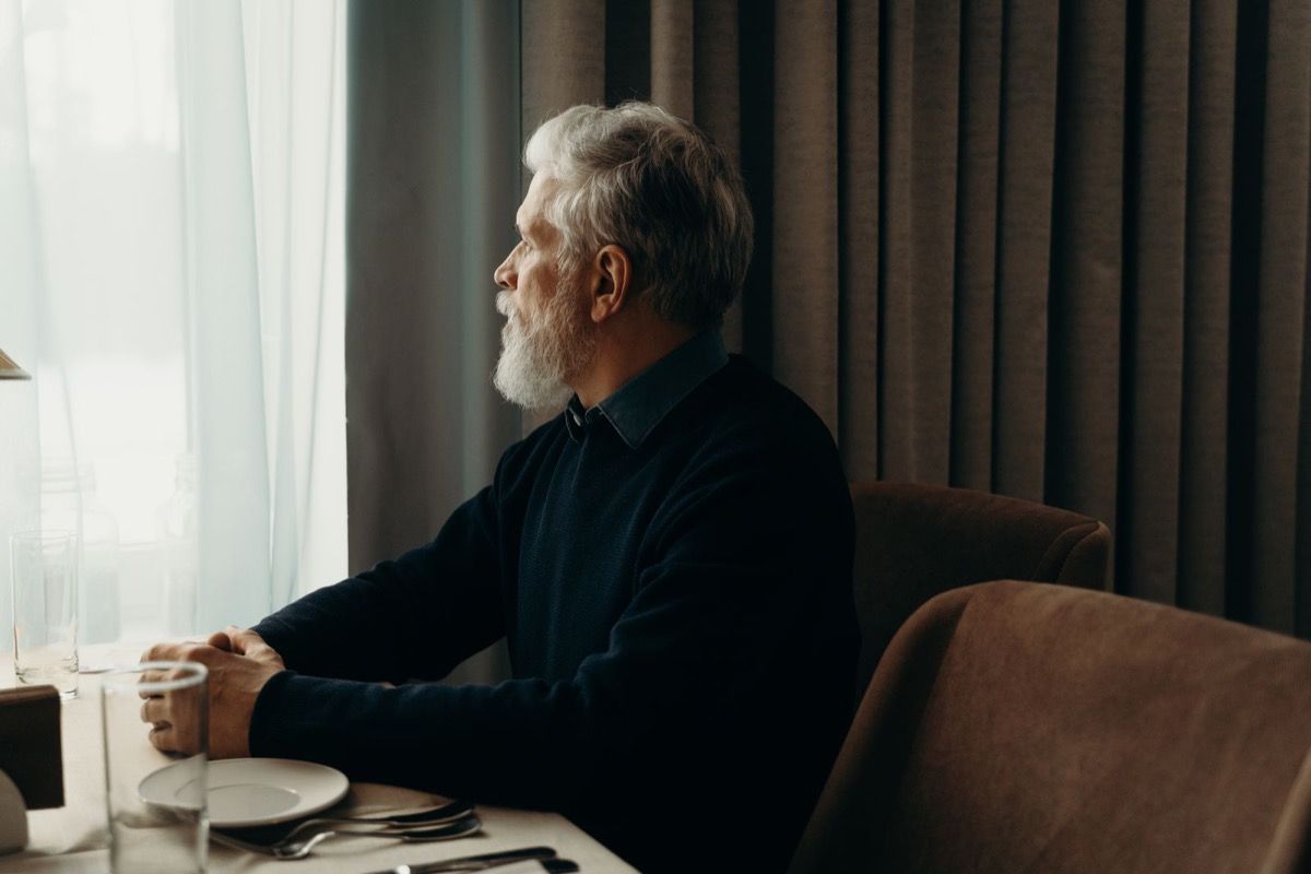 Man over 50 sitting alone at a cafe table with coffee, looking out the window in soft natural light