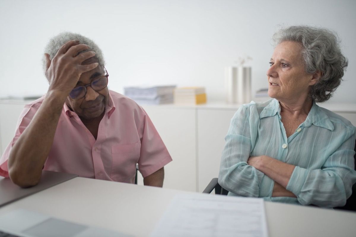 Two older adults seated at a table, one with a hand on his forehead and the other looking toward him